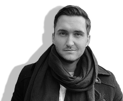 A black and white photograph of artist Mike Emilio. He has short light hair and is wearing a dark jacket with a scarf.