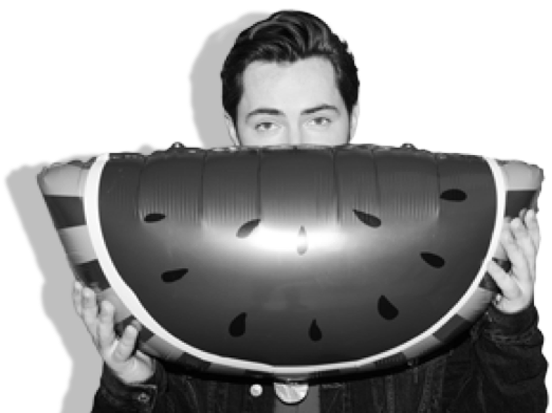 A black and white photograph of artist Steve Void. He has short dark hair and is wearing a dark jacket, and is holding up a large inflatable watermelon which covers up his face below his eyes.